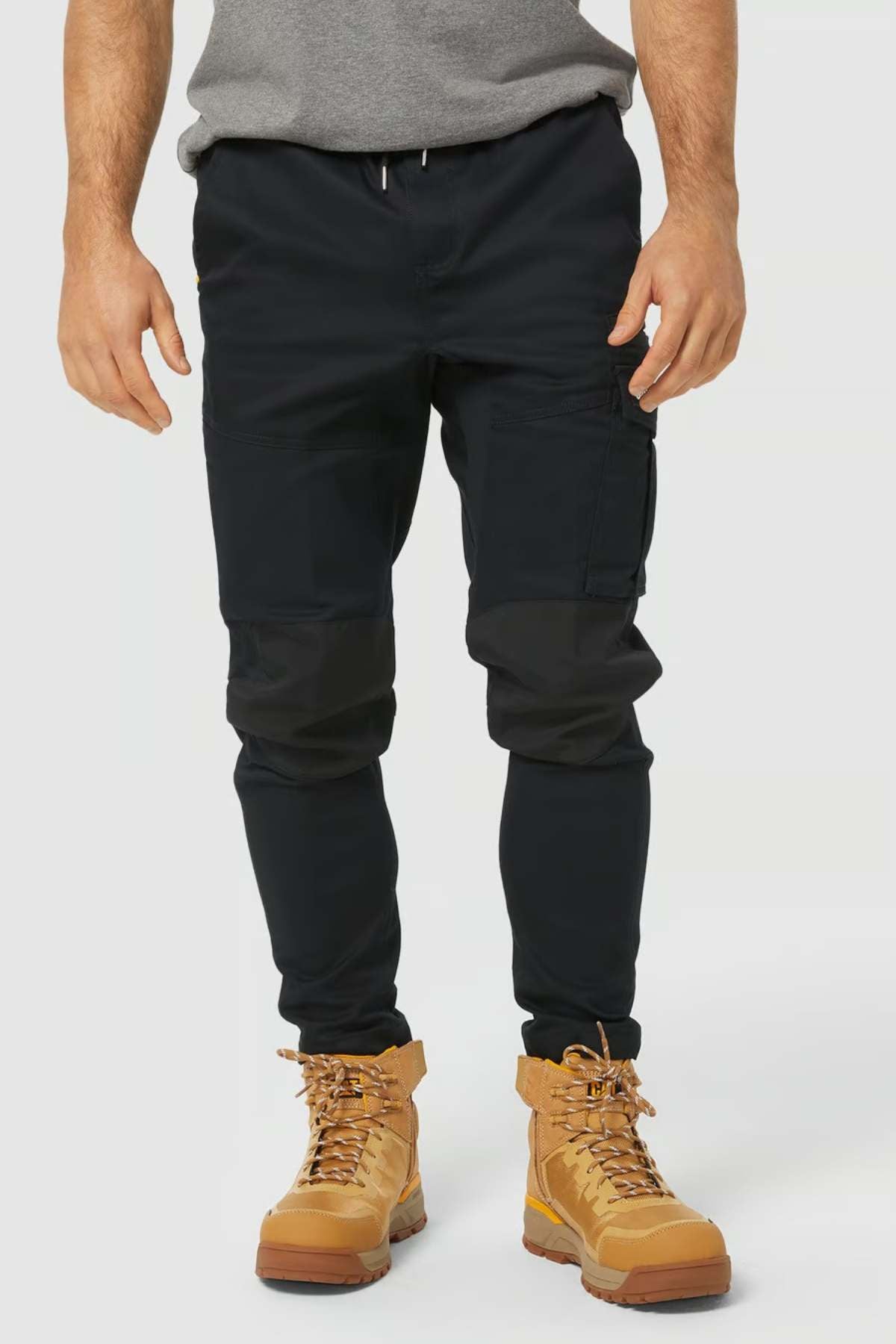 A person wears the CAT Dynamic Slim Fit Pant in black, featuring reinforced knee patches, paired with a gray shirt and tan lace-up work boots. The photo is cropped from the shoulders down against a plain light background.