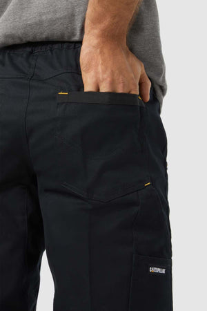 A person in CAT Dynamic Slim Fit Pants with reinforced knee patches and a gray shirt stands with their right hand in the back pocket, showcasing the pants' detailed stitching and functional pocket design.
