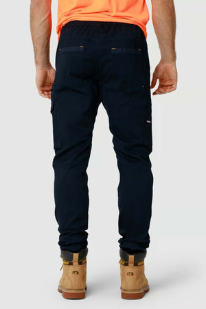 The person is wearing CAT Dynamic Slim Fit black cargo pants with reinforced knee patches, tan work boots featuring black accents, and a bright orange shirt, all against a plain background.