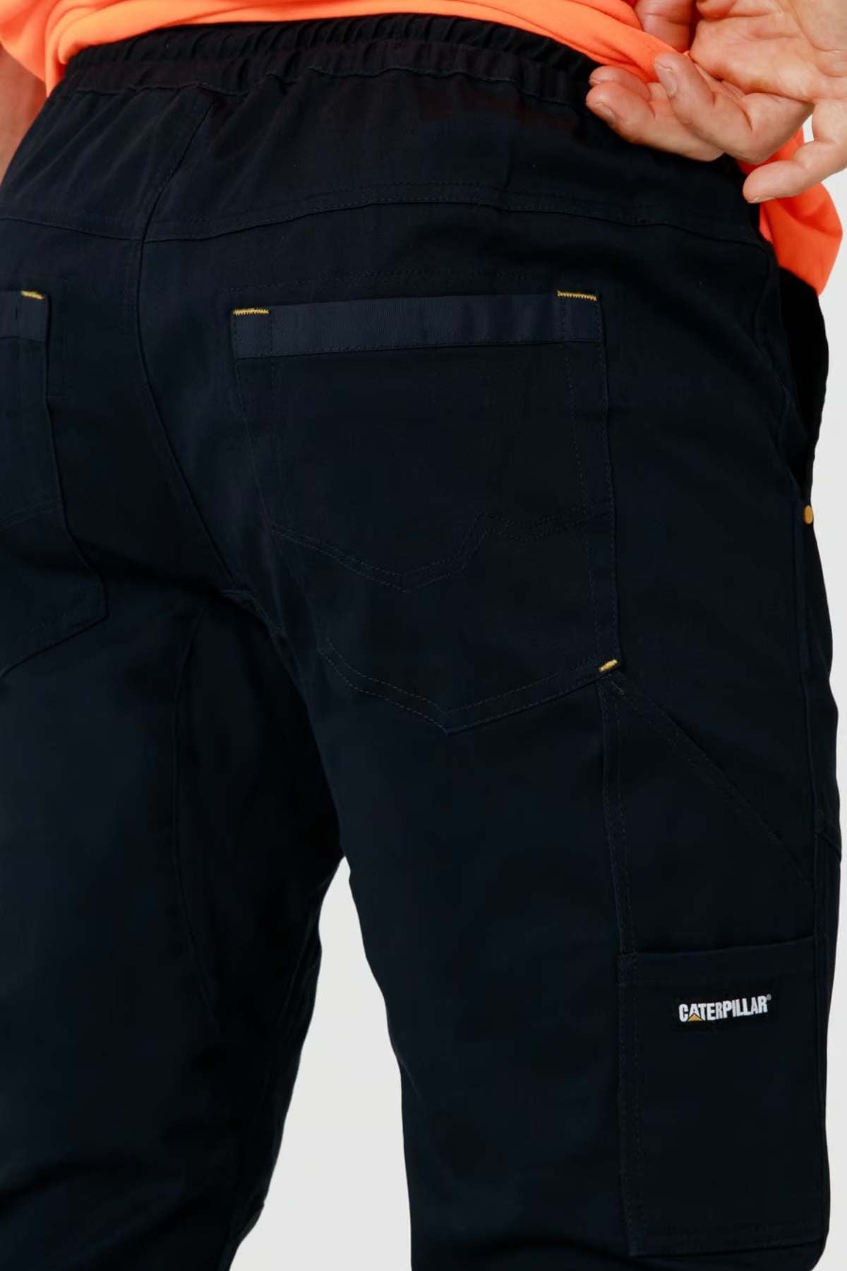 Close-up of a person wearing black CAT Dynamic Slim Fit Pant with visible back pockets and a "Caterpillar" logo on the side pocket; part of an orange shirt is also visible.