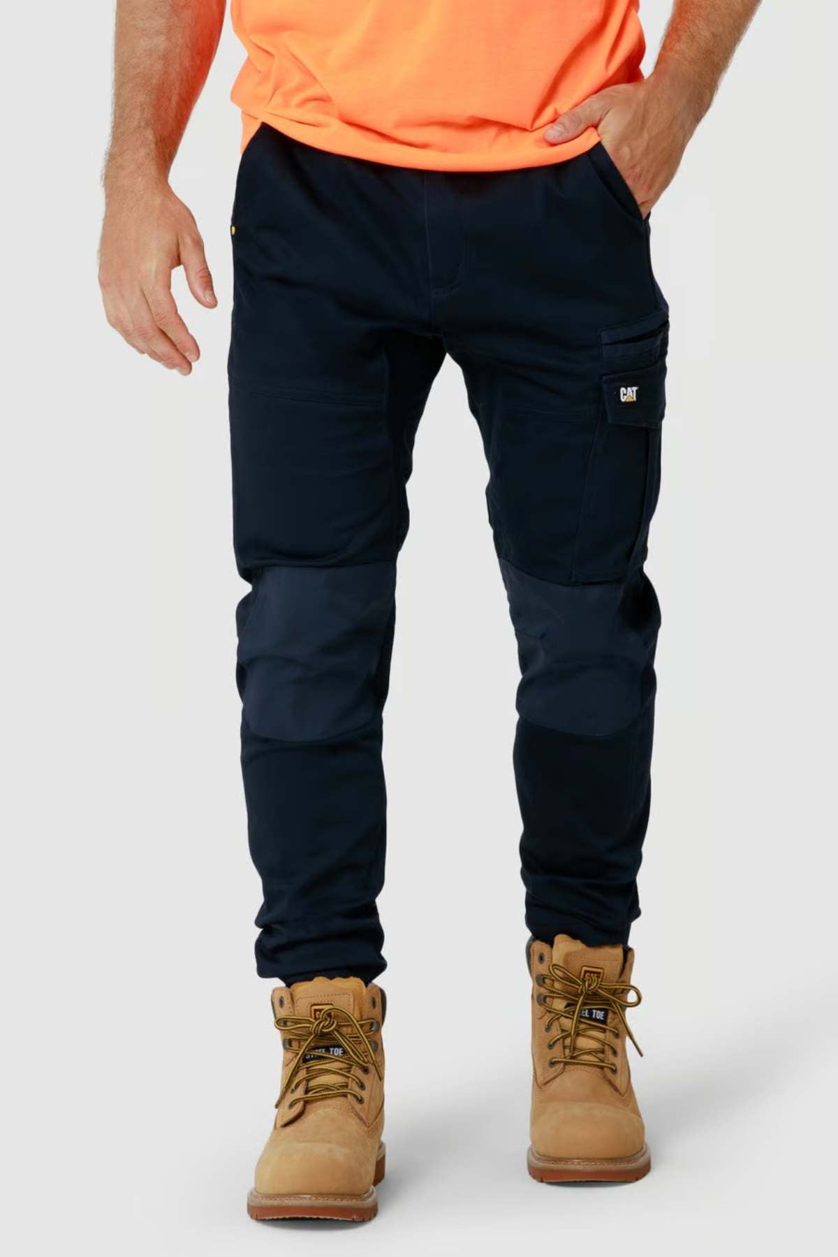 A person stands with one hand in their pocket against a plain light background, wearing a bright orange shirt, tan work boots, and navy CAT Dynamic Slim Fit Pant cargo pants with reinforced knee patches.