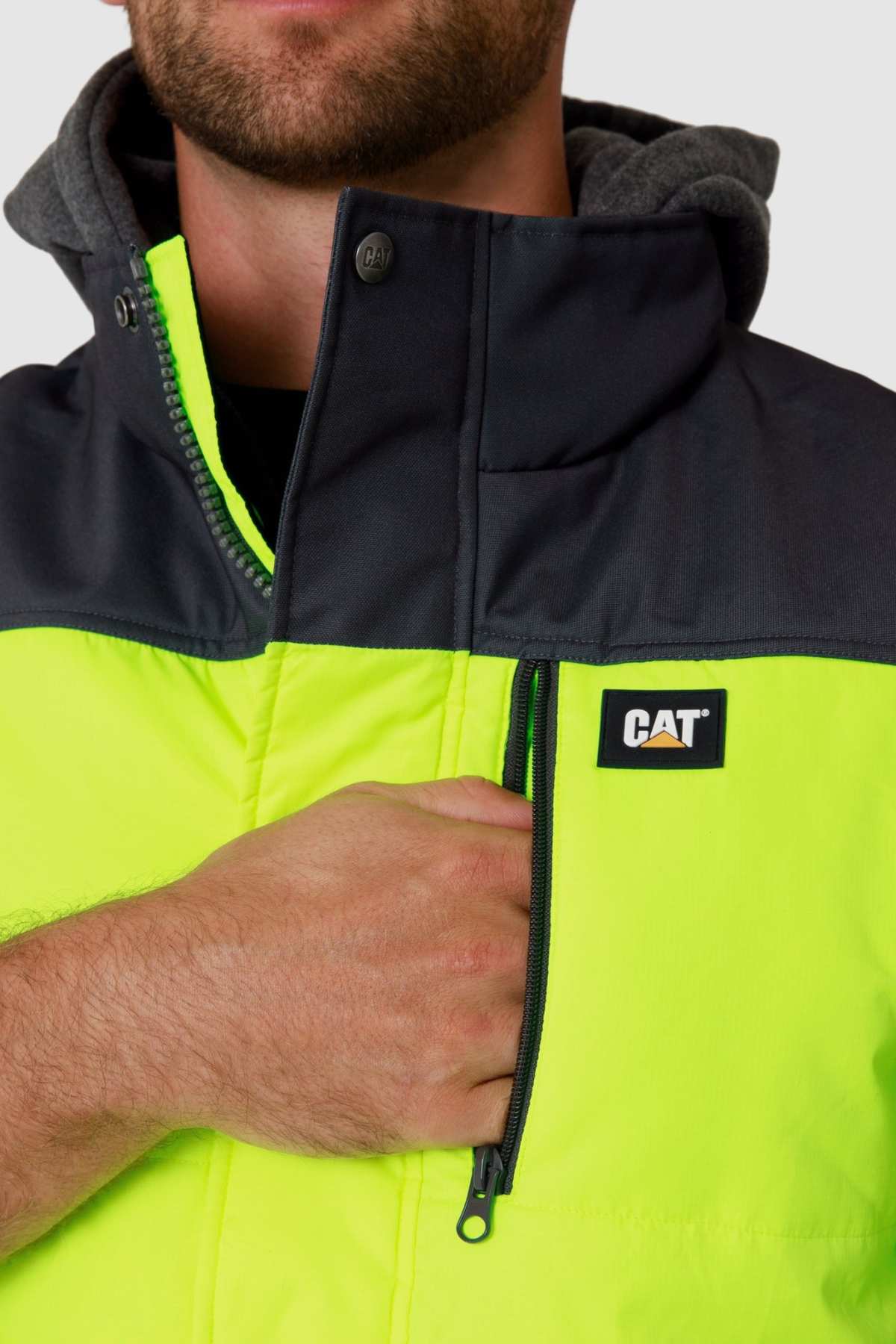 A person wearing a CAT Hi Vis Hooded Work Vest in bright yellow and black is gripping its zipper. A high collar enhances its design, partially obscuring the face while highlighting the water-resistant functionality.