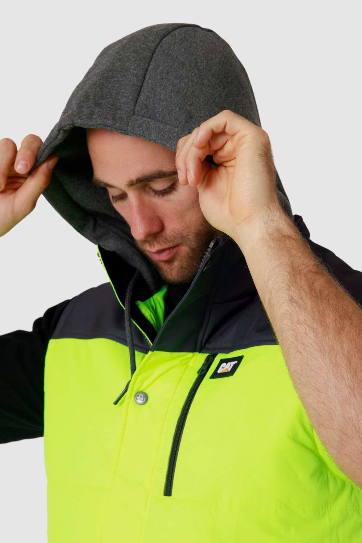 A person adjusts their hood while wearing the CAT Hi Vis Hooded Work Vest in neon green and black with a gray hood. This water-resistant garment features a chest logo, and the person is looking down against a plain gray background.