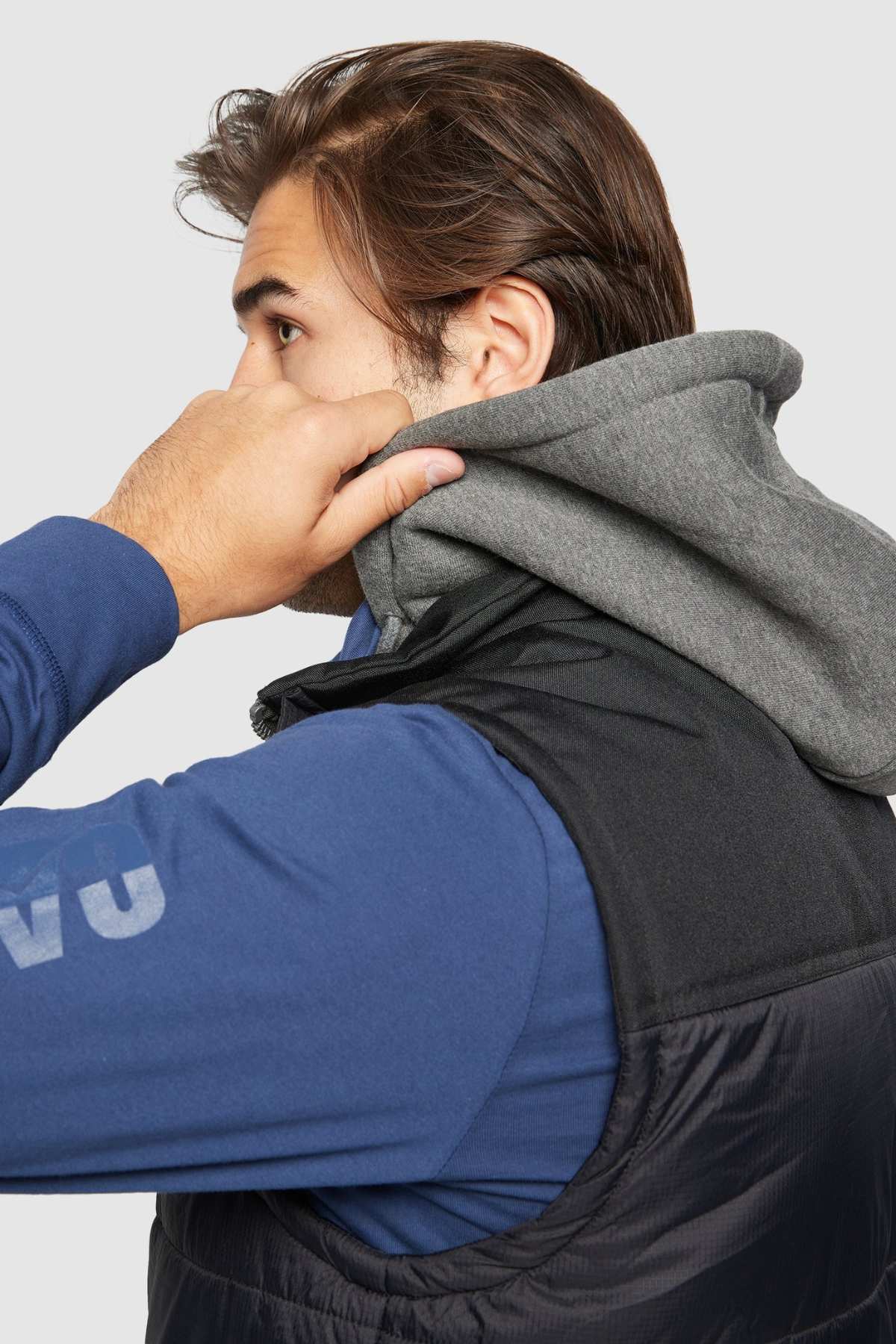 A person with medium-length brown hair is adjusting the hood on their grey hooded sweatshirt. Over the sweatshirt, they are wearing a black CAT Hooded Work Vest, which has multiple pockets and is water-resistant. The background is plain and light-colored, and the person's face is partially obscured as they are turned to the side.