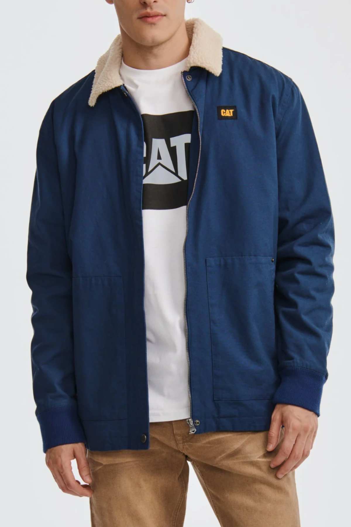 Someone is wearing a blue CAT Sherpa Lined Chore Jacket with a light shearling collar over a white Caterpillar logo t-shirt, paired with tan pants. The jacket features a small yellow and black patch on the chest against a plain white background.