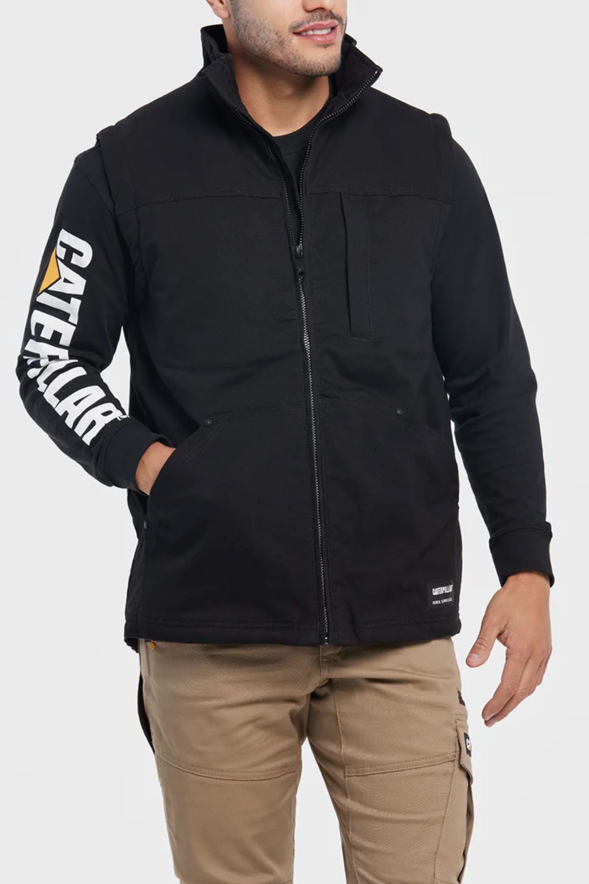 A man wearing a black CAT Sherpa Lined Vest with white "CATERPILLAR" text on the sleeve, beige cargo pants, and his hands in the vest pockets stands against a plain light background.