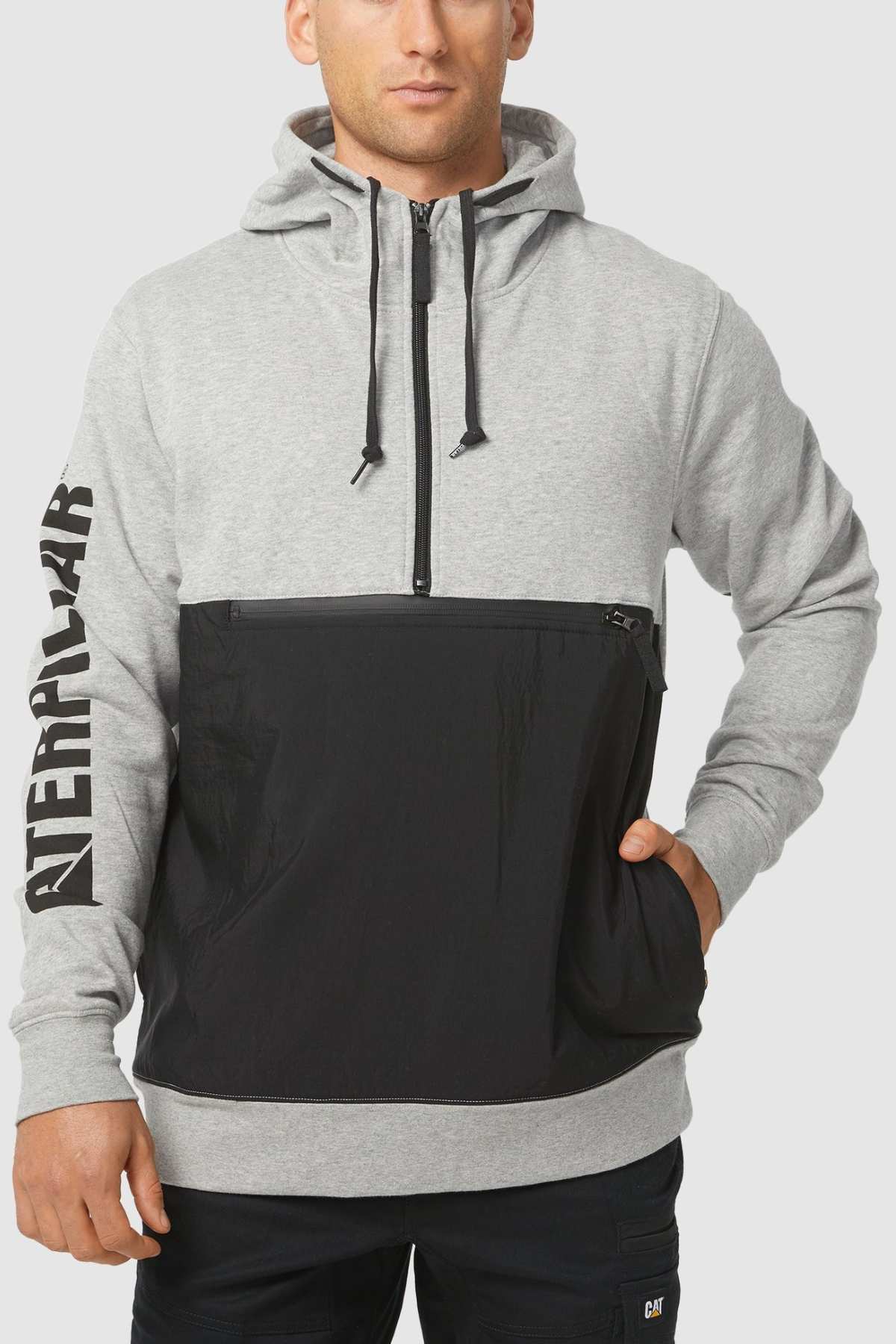 A person in a gray CAT Stillwater 1/4 Zip Hoodie, with "CATERPILLAR" text on the left sleeve and a front black pocket, is posing stylishly with one hand tucked into the pocket.
