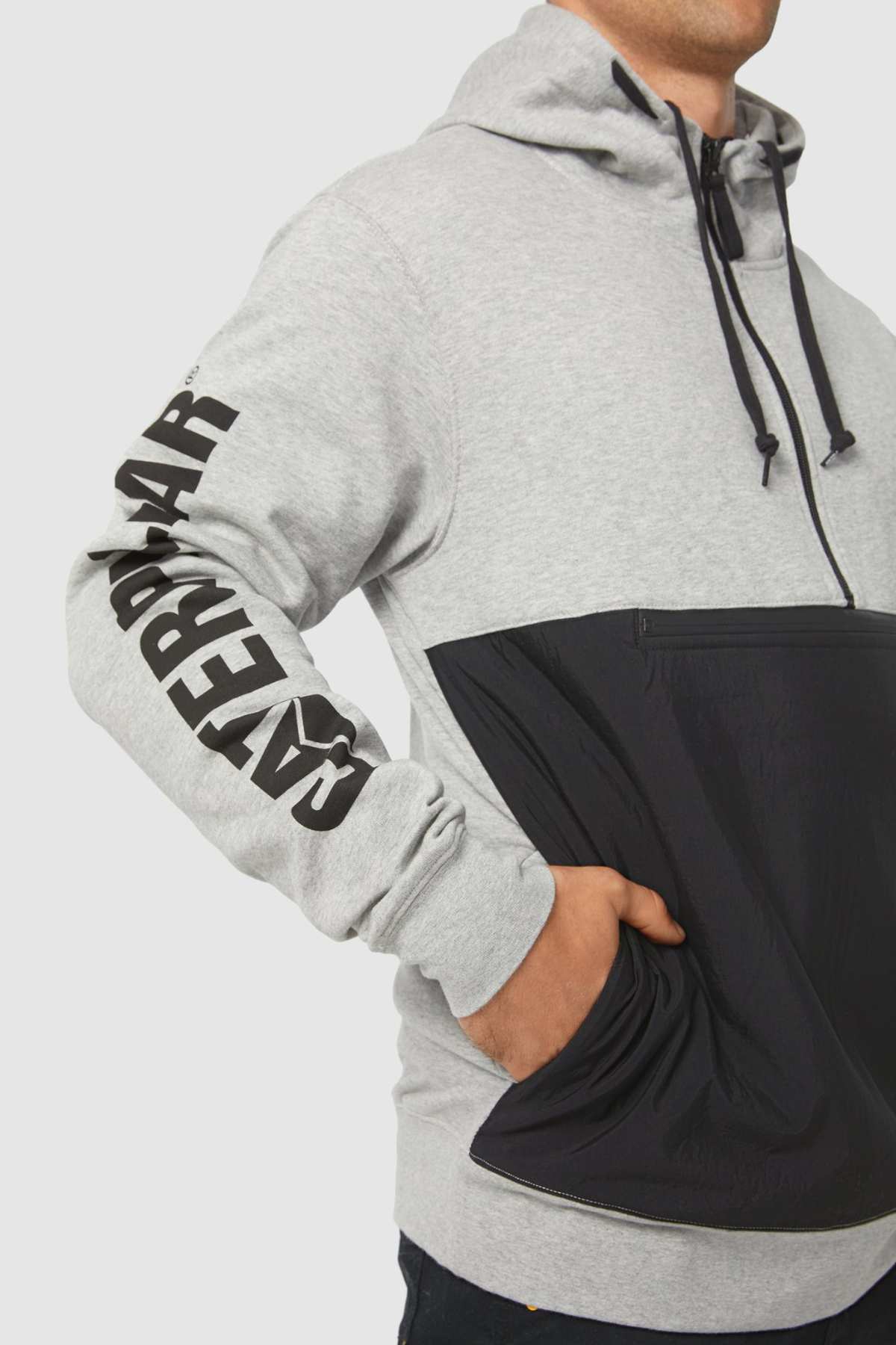 A person in a gray and black CAT Stillwater 1/4 Zip Hoodie with "CATERPILLAR" on the sleeve stands casually with a hand in the front pocket. The hoodie includes a drawstring hood and S3 Cord Management for convenience.