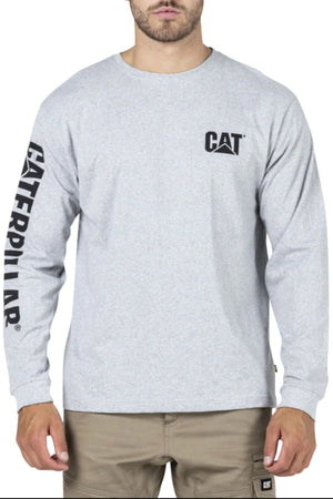 A man wears a CAT Trademark Banner Long Sleeve Tee featuring the CAT logo on the chest and "CATERPILLAR" down the left sleeve, paired with khaki pants, and stands facing forward.
