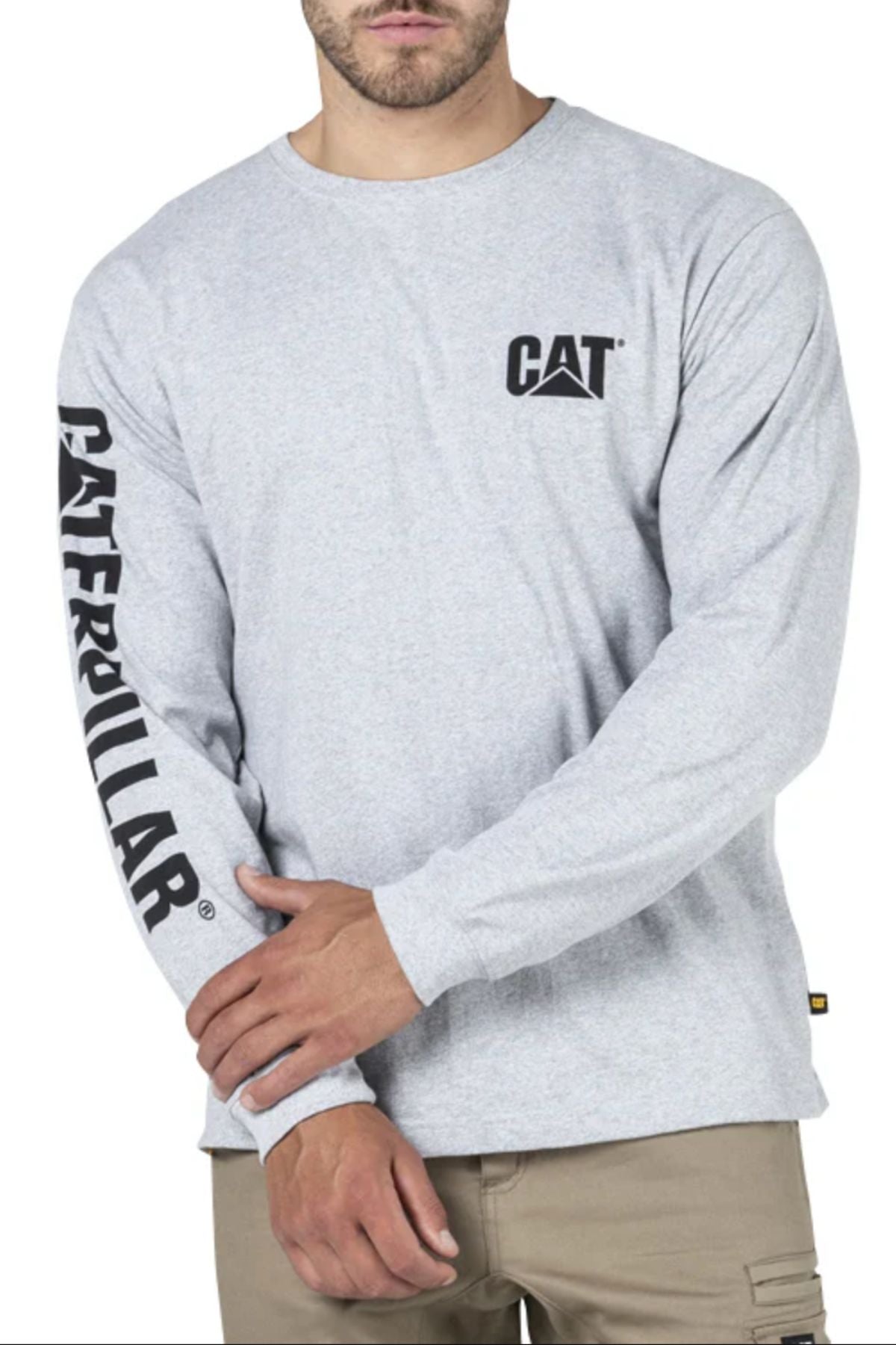 A man wears the CAT Trademark Banner Long Sleeve Tee in light gray, featuring a "CAT" logo on the chest and "CATERPILLAR" on the right sleeve, paired with tan pants for a comfortable, relaxed everyday look.