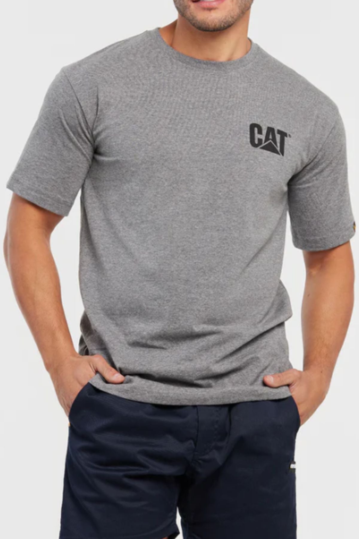 A man stands against a plain background wearing a gray CAT Trademark Tee with a crew neck and small black "CAT" logo on the left chest, paired with navy blue shorts and his hands in his pockets.
