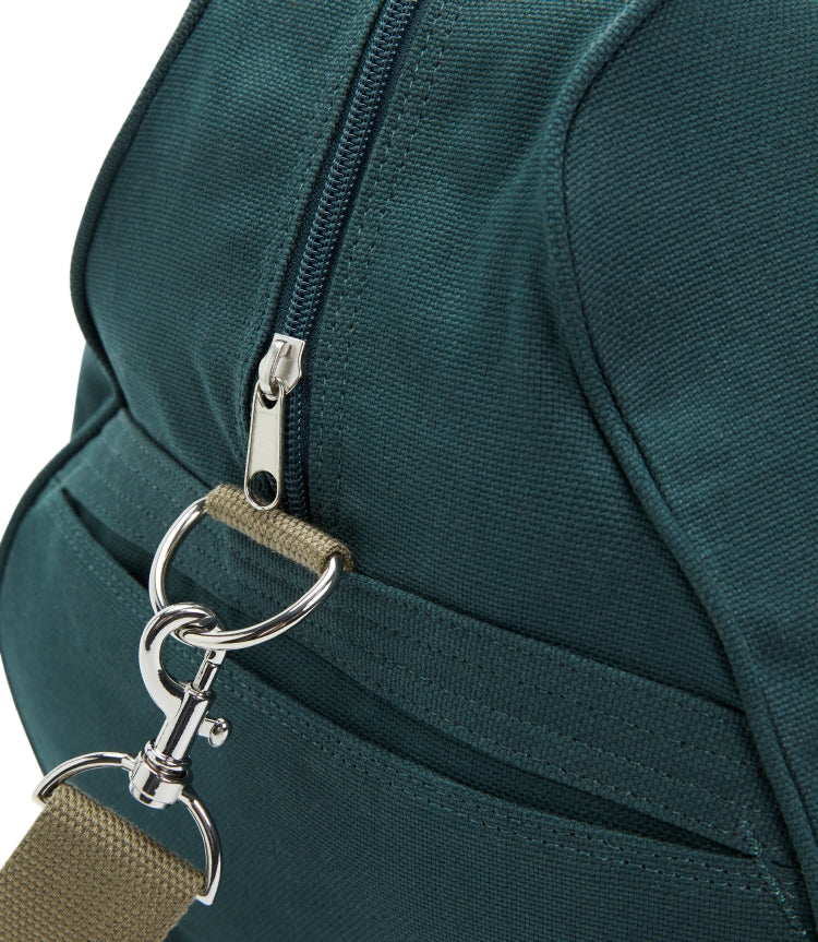 Close-up of the RM Williams Nanga Canvas Bag, a duffle-style accessory made from durable cotton canvas, featuring teal fabric, a silver zipper, metal clasp, beige strap, and detailed stitching.