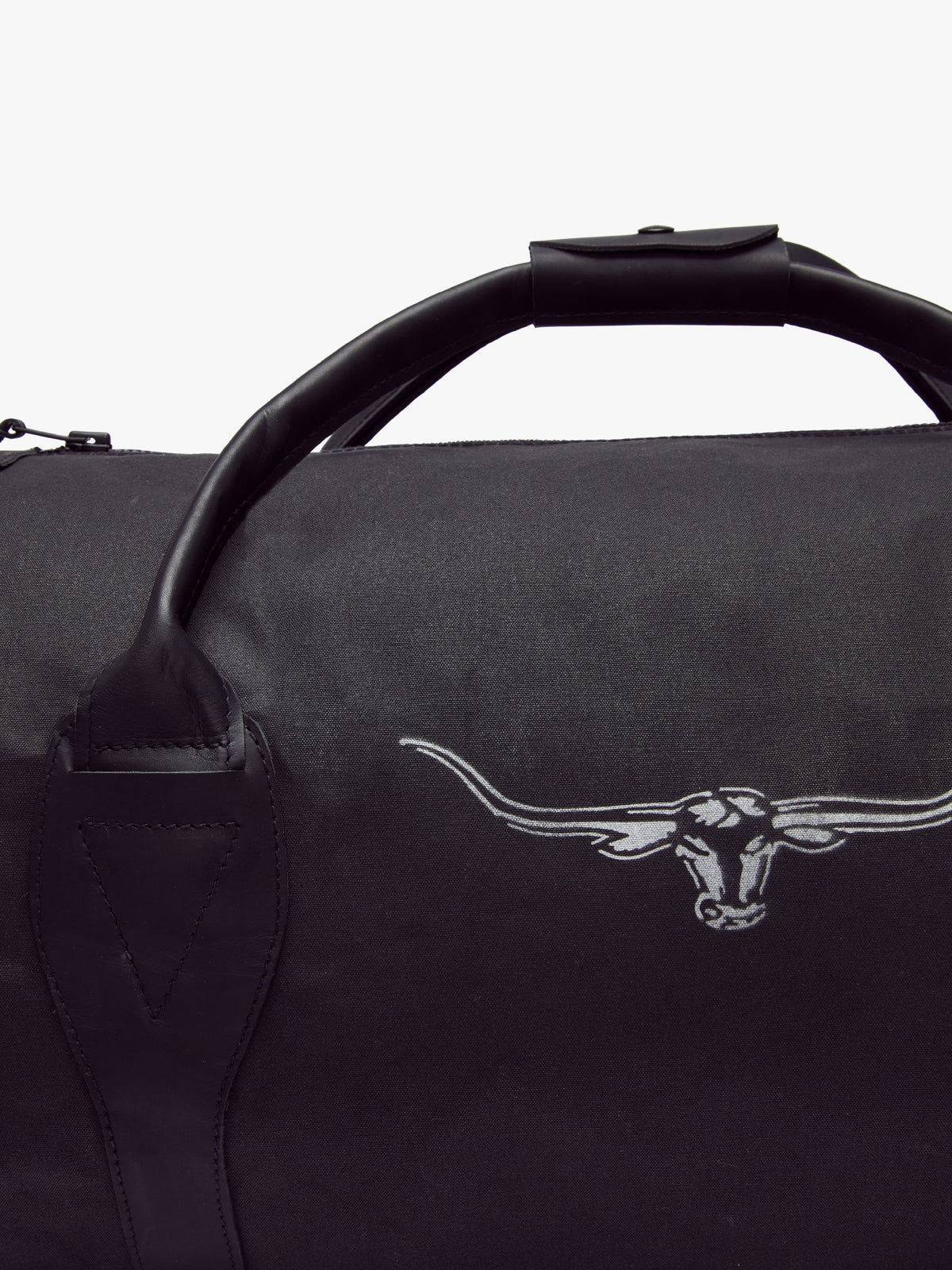 The RM Williams Canvas Ute Bag is a black duffel with water-resistant fabric, leather handles, and a white longhorn steer head outline. The image shows part of the handle and the left side of the bag.