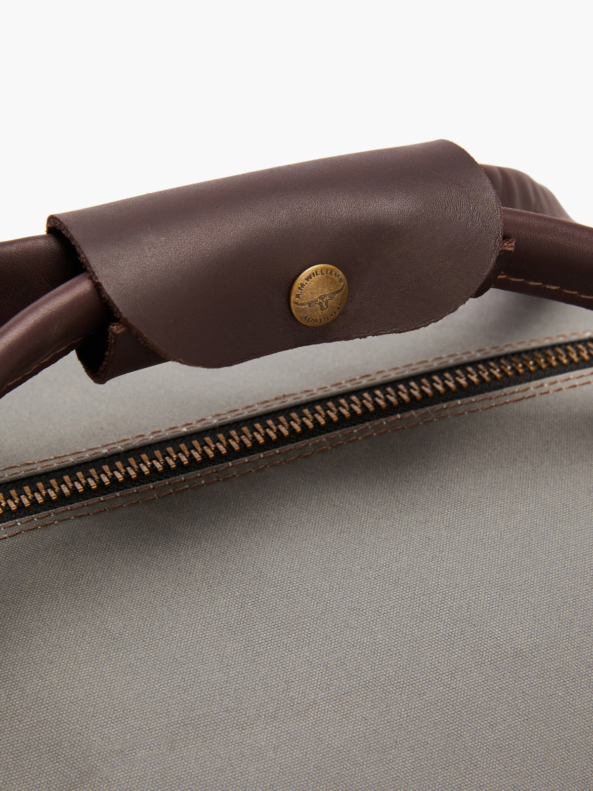 Close-up of the RM Williams Canvas Ute Bag highlighting a dark brown leather handle with a "Made in USA" brass button. The water-resistant light gray utility canvas features a visible brown zipper and tan stitching.