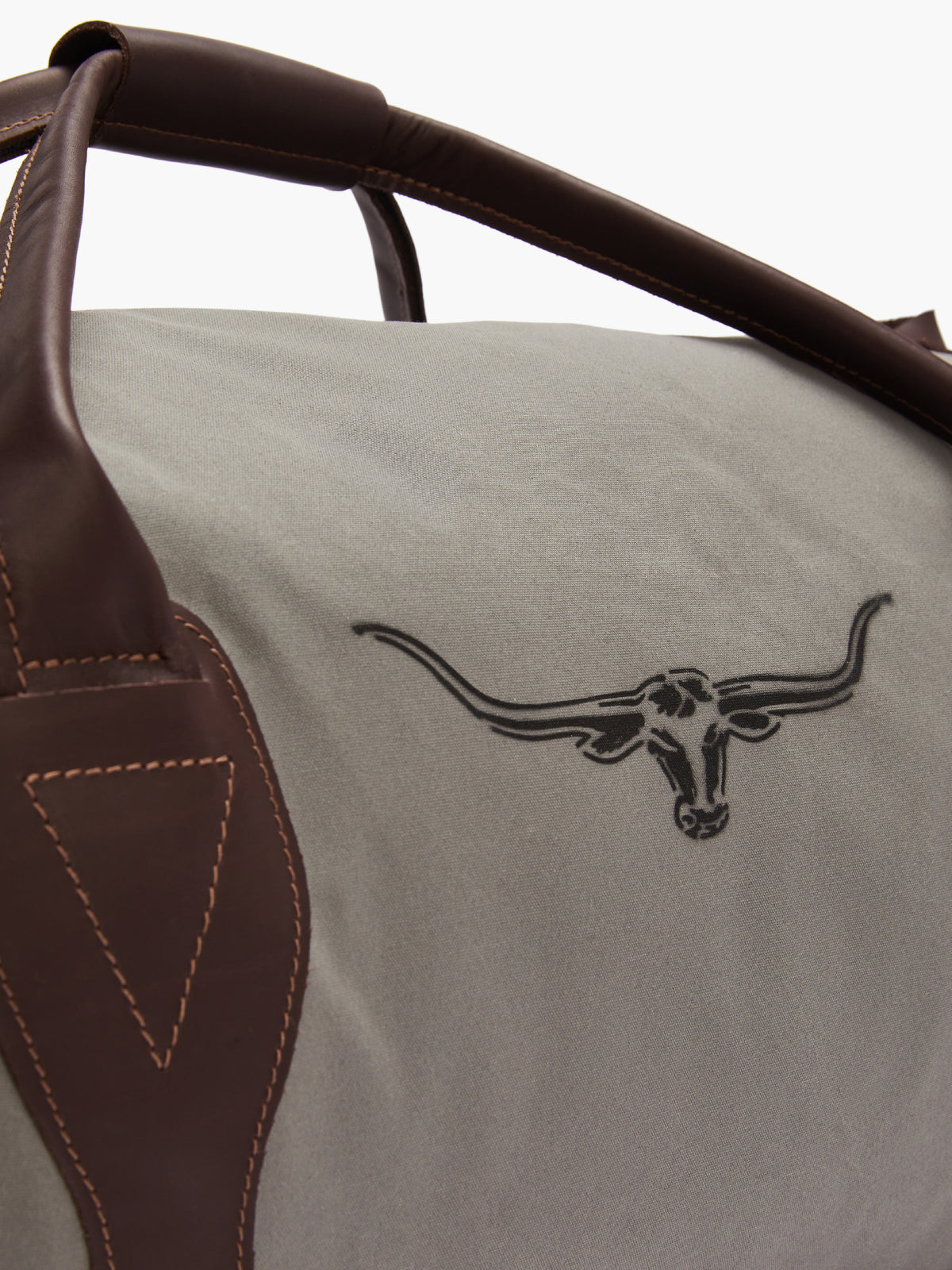 Close-up of the RM Williams Canvas Ute Bag's corner, featuring durable gray utility canvas with a dark longhorn cattle logo. The water-resistant bag includes dark brown leather straps with prominent stitching.