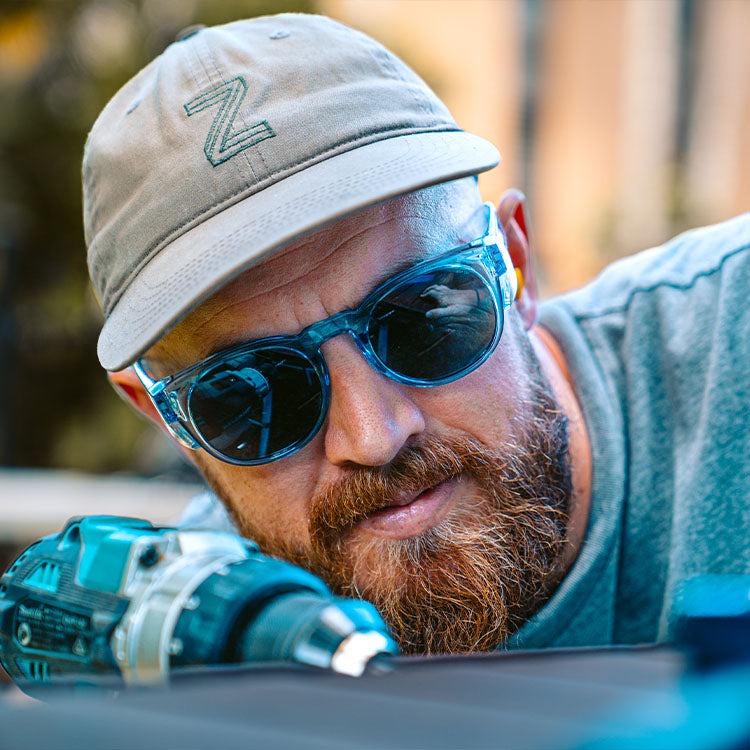 A bearded man in a cap and SafeStyle Cruisers Blue Frame Tinted Lens sunglasses closely examines a power drill, focusing intently on his outdoor work.