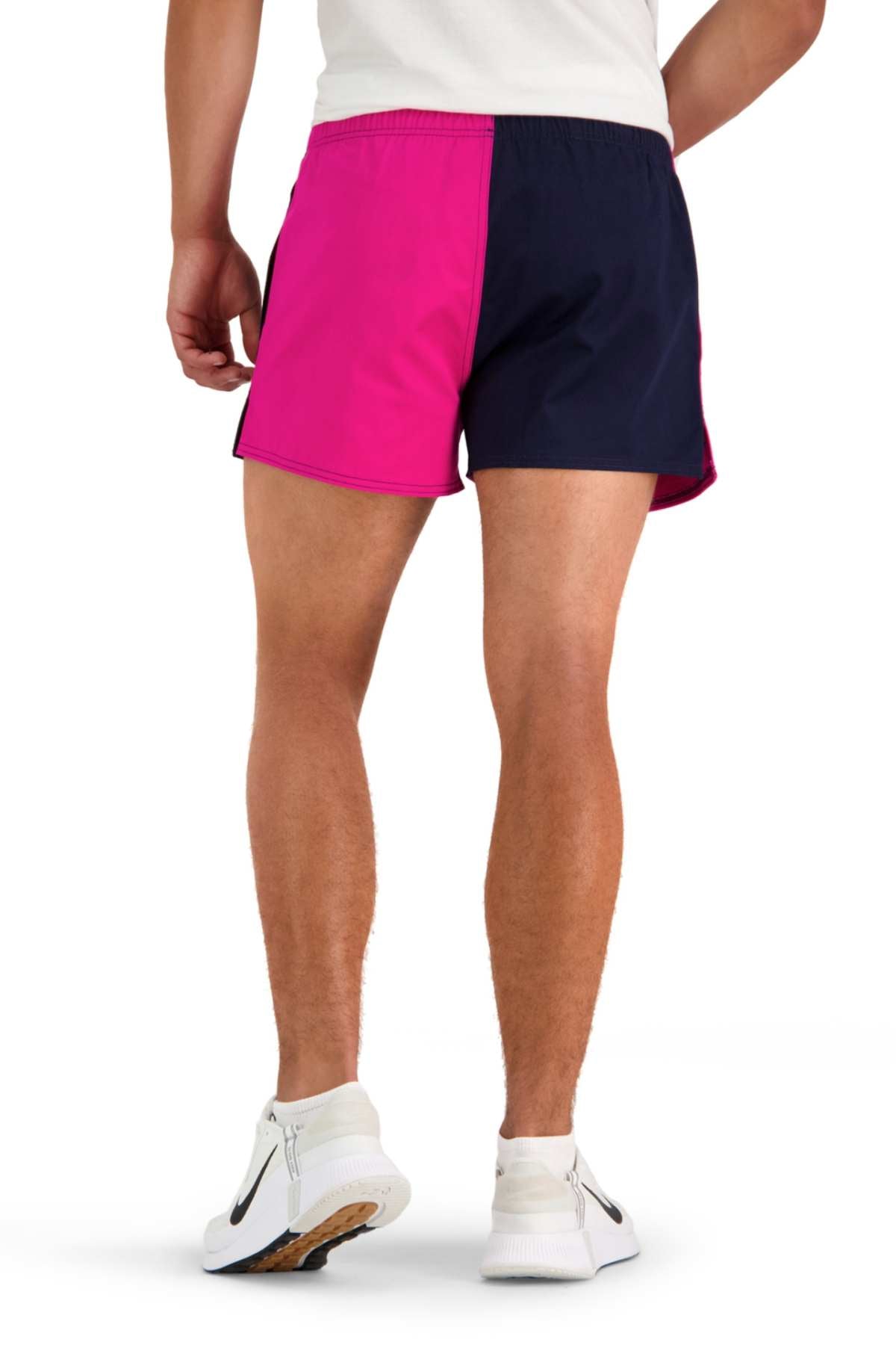 Rear view of an individual standing, donning the Canterbury Cotton Twill Harlequin Short in a two-tone design with one side pink and the other navy blue. They are also wearing white athletic shoes adorned with black and orange accents, providing breathable comfort for any activity.