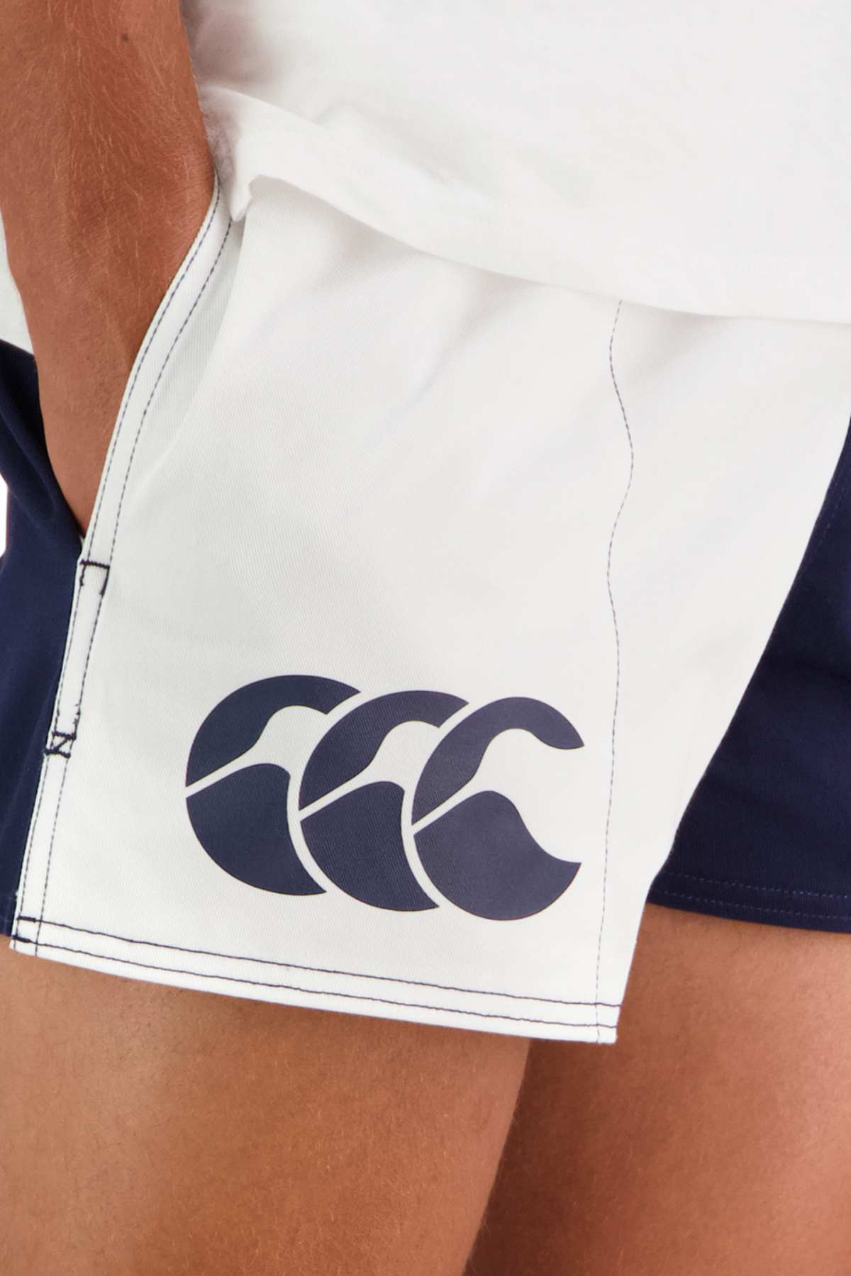 Close-up of a person wearing the Canterbury Cotton Twill Harlequin Short, white athletic shorts with blue accents. Known for their breathable comfort and customizable fit, these shorts feature a navy blue logo with three stylized waves on the left side. The person's hand is resting near the pocket of the shorts.