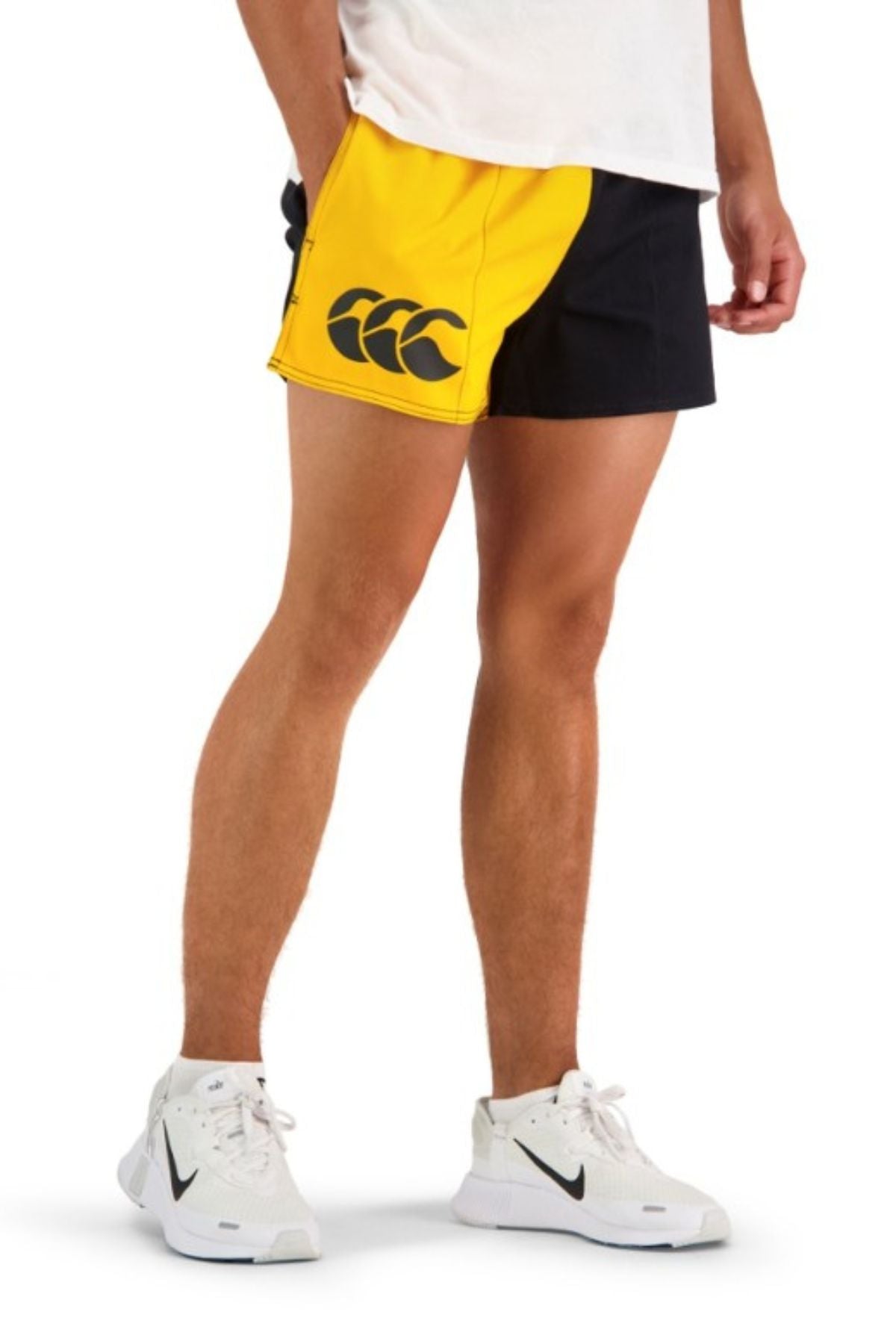 A person wearing a white shirt, Canterbury Cotton Twill Harlequin Shorts with an elasticated waist, and white Nike sneakers stands with one hand in their pocket against a plain background.