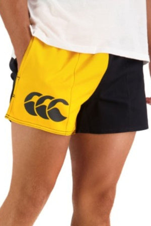 A person wears the yellow and black Canterbury Cotton Twill Harlequin Short, featuring an elasticated waist and a large left-side logo, paired with a white shirt. Shown from mid-torso to mid-thigh.