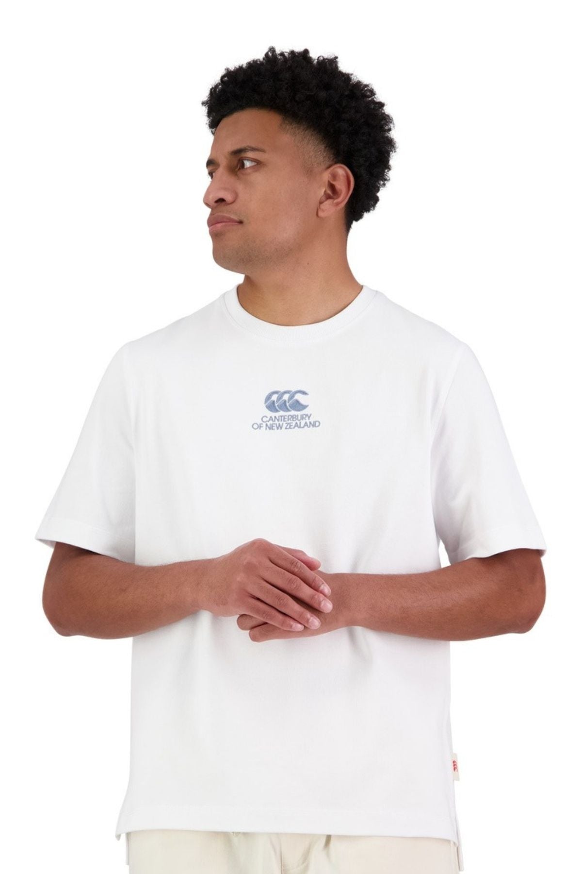 A man wearing the Canterbury Heritage T-Shirt—an oversized, heavy-duty 100% cotton white tee with a small blue CCC logo on the chest—stands against a white background, looking to his left with his hands loosely clasped.