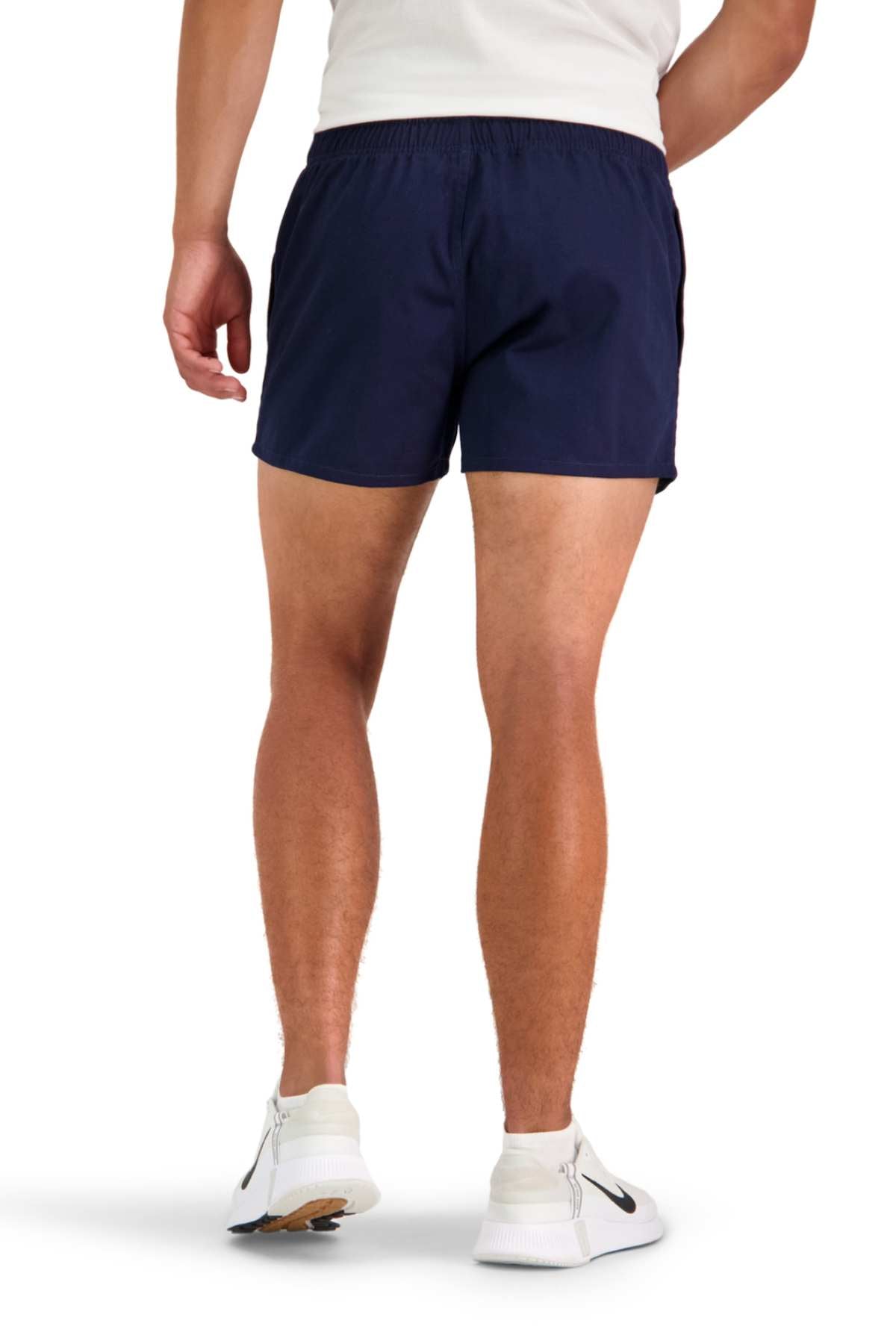 A person is seen from behind, wearing a white shirt and navy blue Canterbury Rugged Drill Short featuring an elasticated waist. They are also sporting white athletic shoes with black accents against a plain white background.