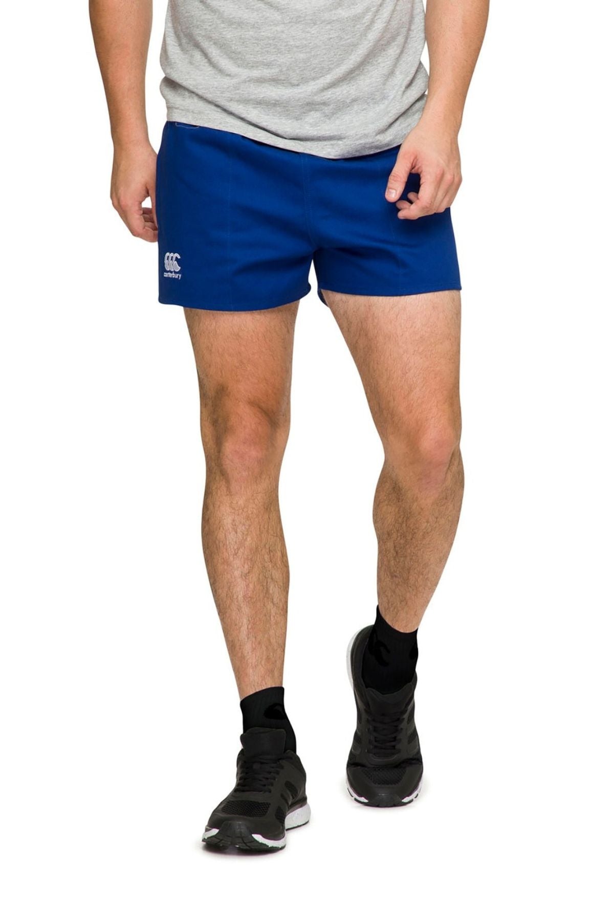 The image showcases a person wearing a gray shirt paired with Canterbury Rugged Drill Shorts in royal blue, featuring the logo and designed with an elasticated waist for optimal comfort. Black socks and black athletic shoes complete the look. The focus is on the lower half, emphasizing the tape side pockets of the shorts.