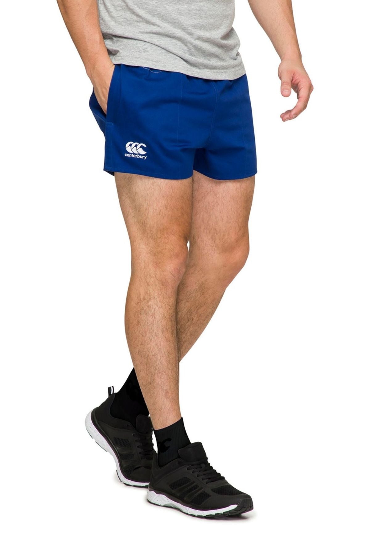 A person wearing a gray T-shirt and blue Canterbury Rugged Drill Short, featuring an elasticated waist, complemented by black socks and athletic shoes, stands against a white background.