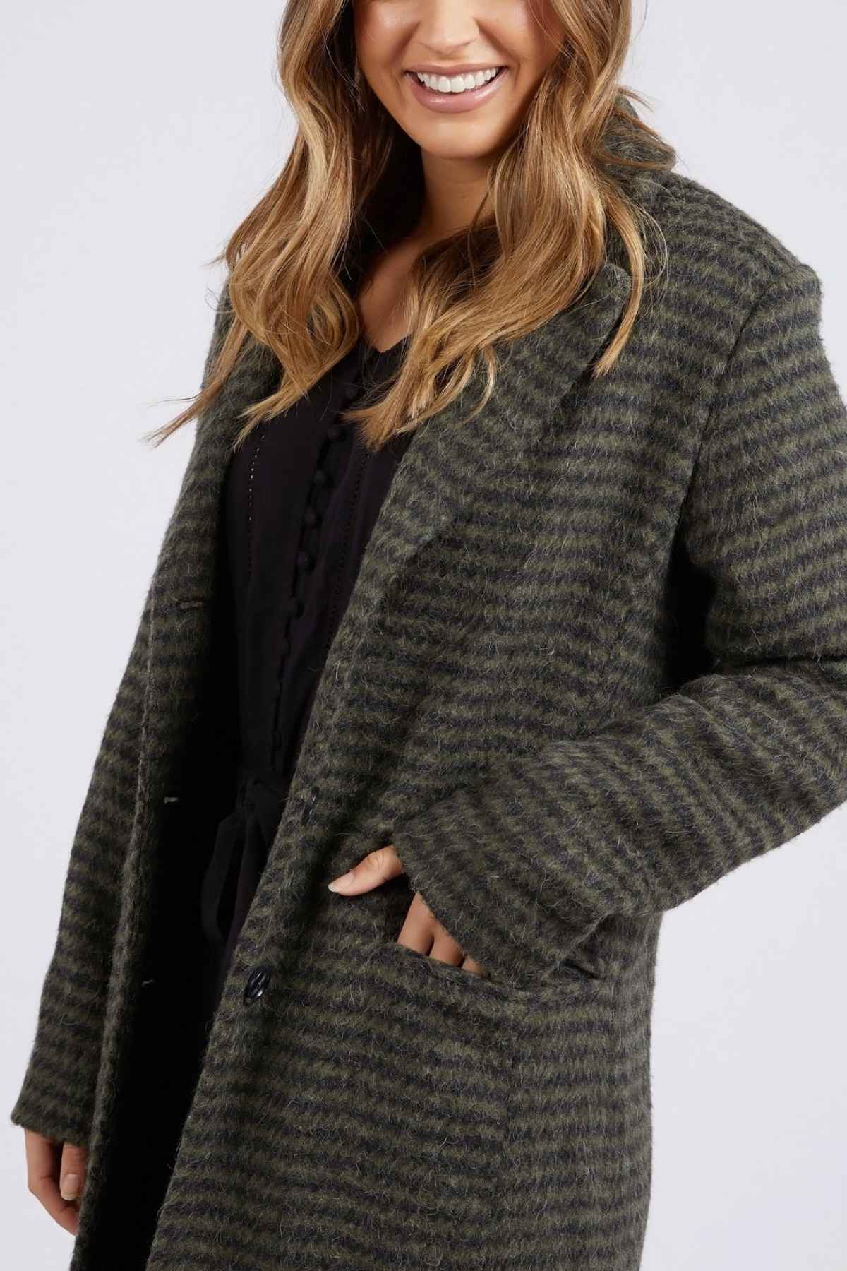 A woman wearing the Foxwood Clementine Coat, a long, grayish-green striped wool blend midi jacket, over a black outfit. She has light brown, wavy hair and is smiling with her right hand in the coat pocket. The elegant jacket adds a refined touch to her appearance against the plain white background.