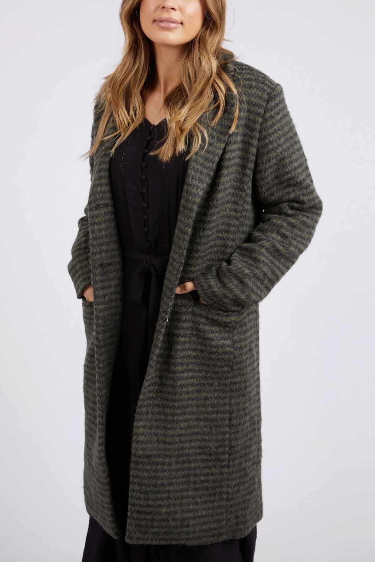A person with long, wavy hair is wearing a dark green Foxwood Clementine Coat over a black outfit. Their hands are tucked into the pockets of the coat as they stand against a plain grey background. The upper part of their face is obscured in the image.
