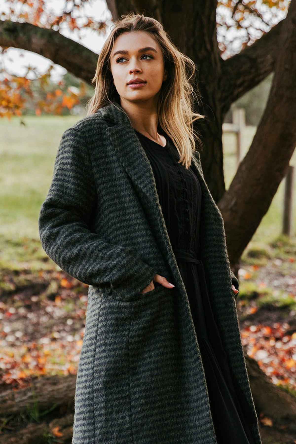 A person with long hair stands outdoors, looking thoughtfully into the distance. They are wearing the Foxwood Clementine Coat featuring a cozy wool blend and a stylish check pattern, with their hands tucked into the coat’s pockets. Autumn leaves and a large tree are visible in the background.
