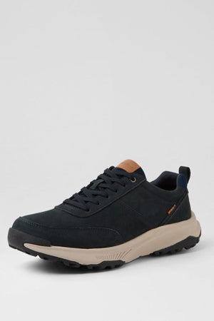The Colorado Suzuki Shoe is a black casual sneaker featuring a beige slip-resistant sole, black laces, a brown tongue accent, and a small orange side tag, shown against a light gray background.