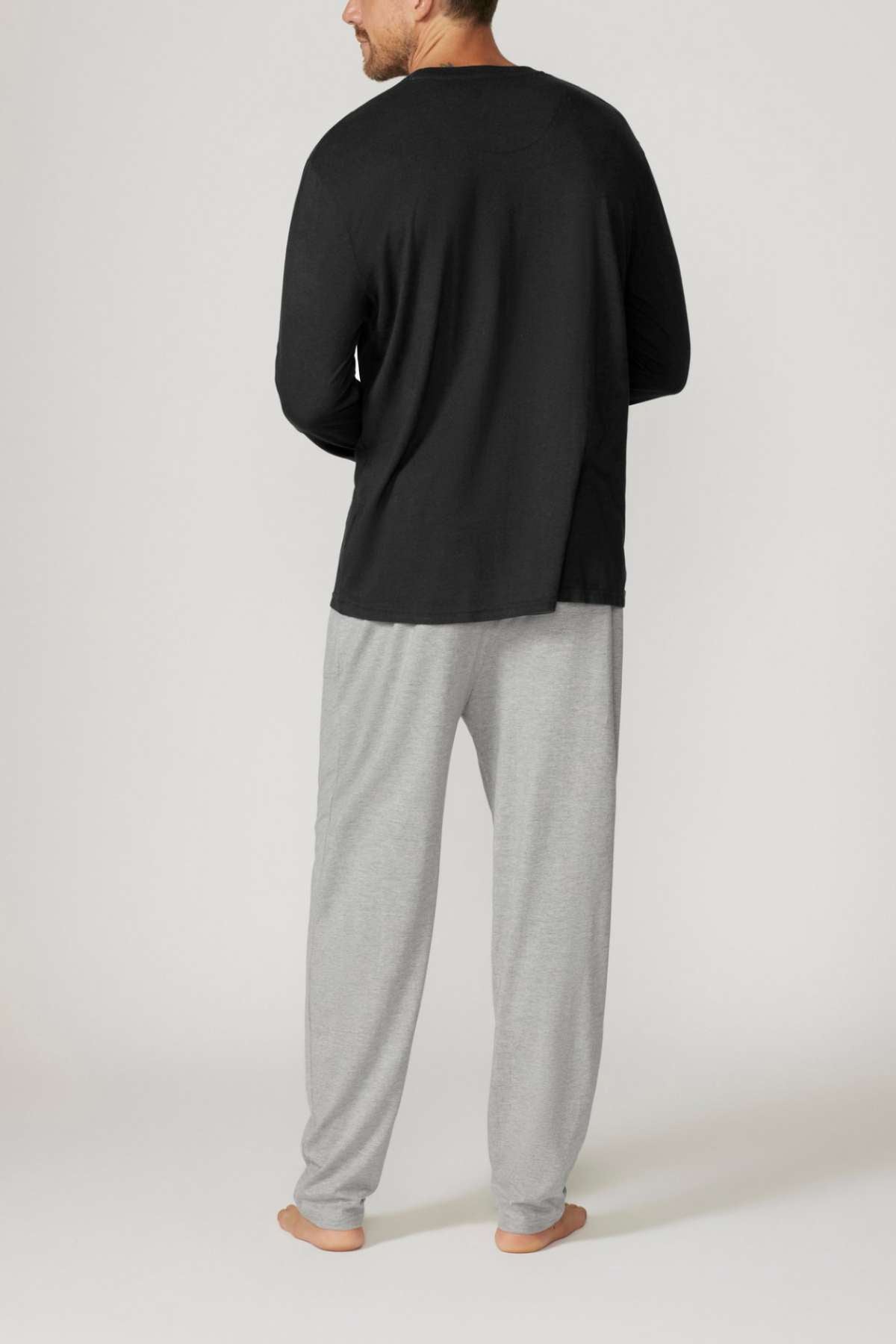 A man stands barefoot in a relaxed fit ensemble, wearing the Contare Country Bamboo Cotton Long Sleeve Set, featuring a black shirt and light gray pants made from hypoallergenic bamboo cotton against a plain, light-colored backdrop.