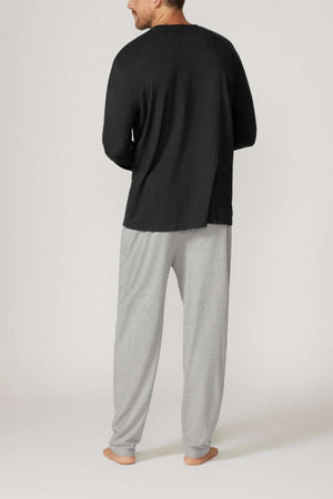 A man stands barefoot in a relaxed fit ensemble, wearing the Contare Country Bamboo Cotton Long Sleeve Set, featuring a black shirt and light gray pants made from hypoallergenic bamboo cotton against a plain, light-colored backdrop.