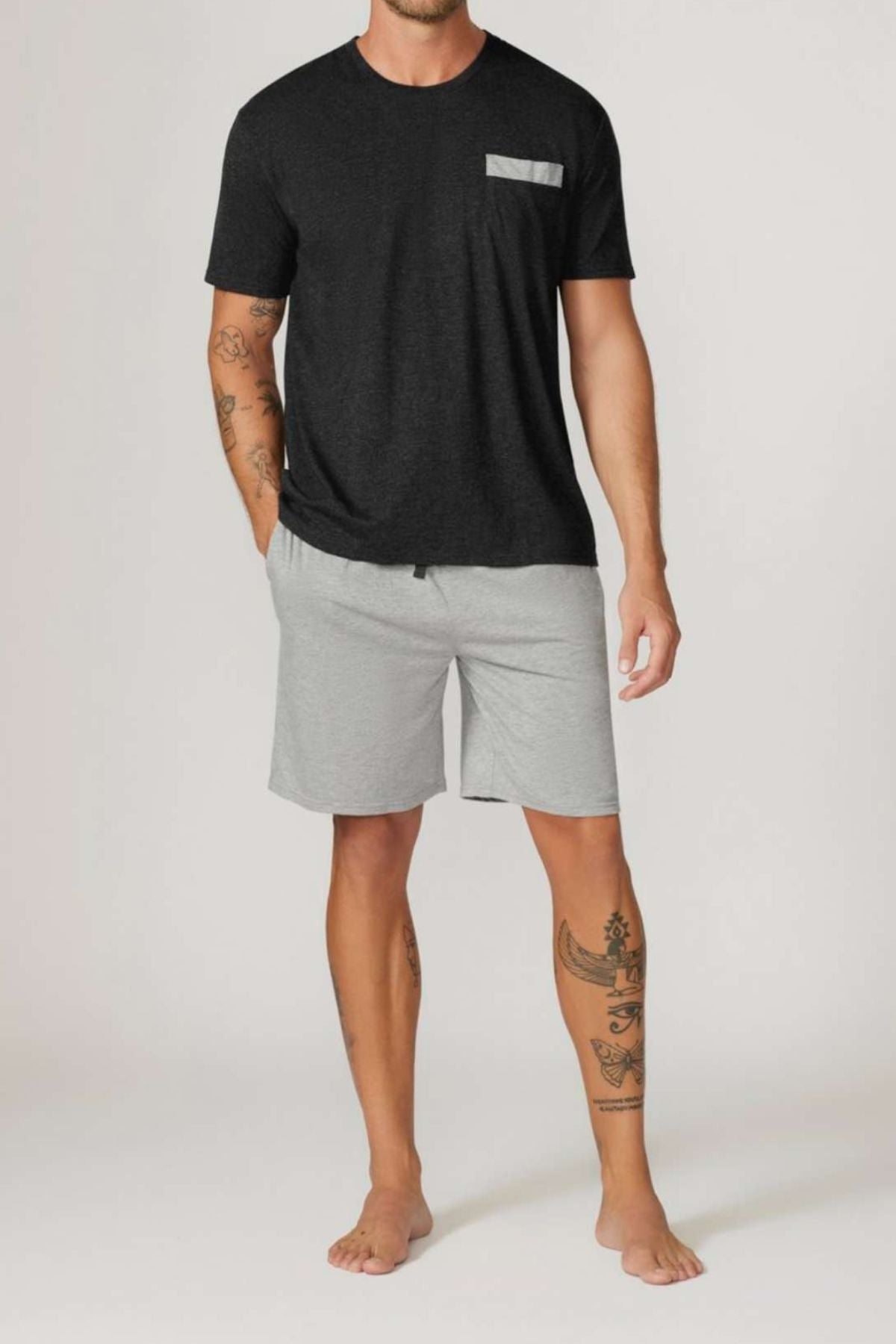 A man with tattoos on both arms and his right leg wears the Contare Country Bamboo Cotton Short Sleeve Set—a black tee with a gray pocket and light gray shorts—standing barefoot against a plain, light background.
