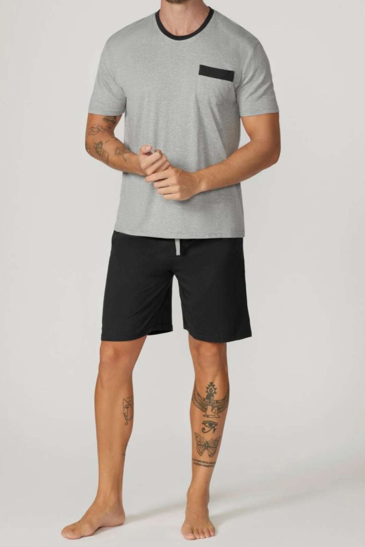 A barefoot man with leg tattoos wears the Contare Country Bamboo Cotton Short Sleeve Set in light grey, featuring a black chest pocket and matching black shorts, standing against a plain background.