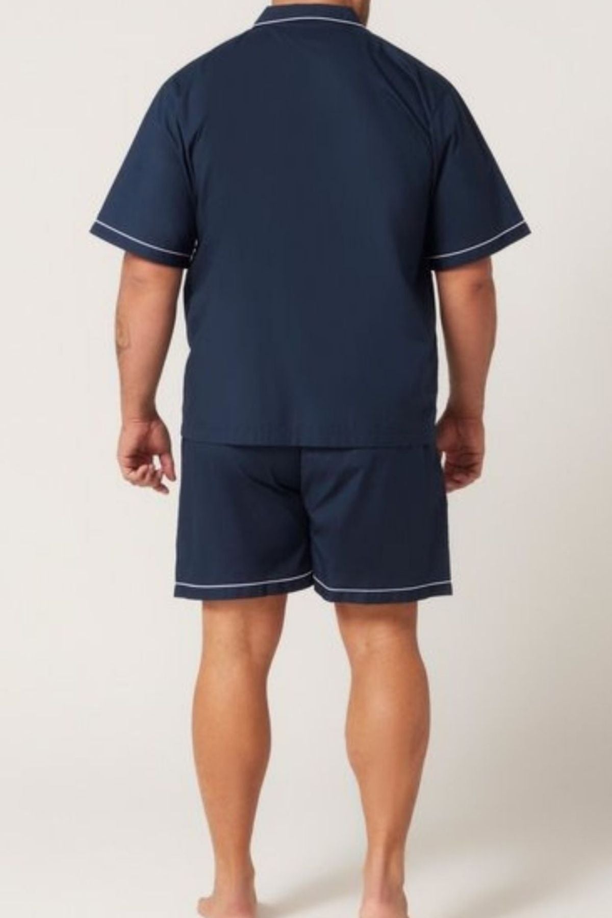 A person stands barefoot, facing away, wearing the Contare Country Cotton Rich Short Sleeve Classic Set in navy with white trim—featuring a short-sleeved top and shorts—against a plain light background.