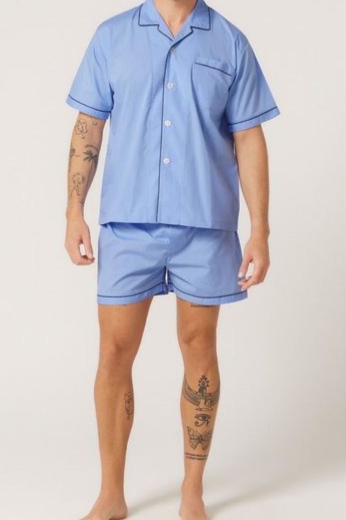 A person with tattoos on their arms and legs is wearing the Contare Country Cotton Rich Short Sleeve Classic Set—light blue pajamas with matching shorts—standing barefoot against a plain light background.