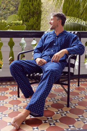 A man wearing the Contare Country Winter Flannelette PJ Set sits barefoot on a tiled balcony surrounded by green plants, looking off to the side and smiling.
