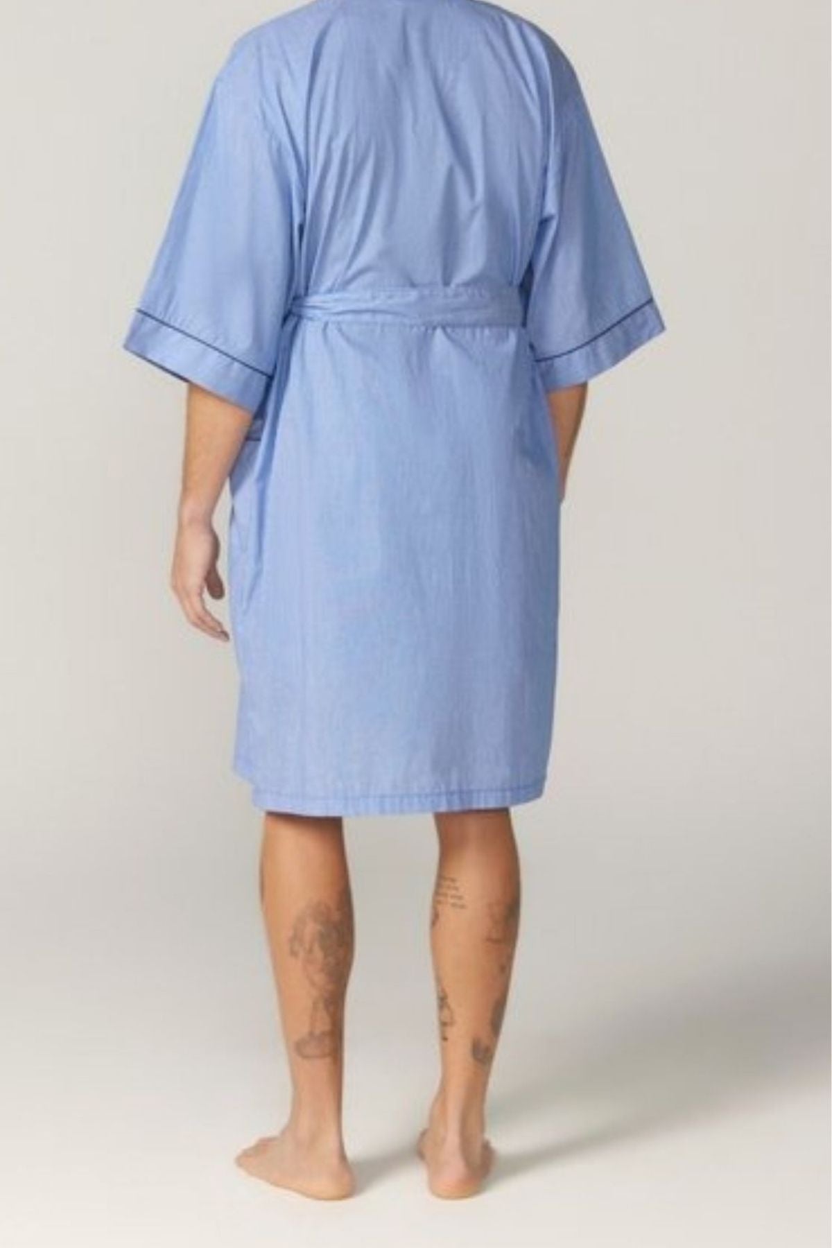 A person stands barefoot, facing away in a light blue Contare Country Summer Dressing Gown. The cotton kimono-style robe has short sleeves, a belt, and shows tattoos on the lower legs against a plain, neutral background.