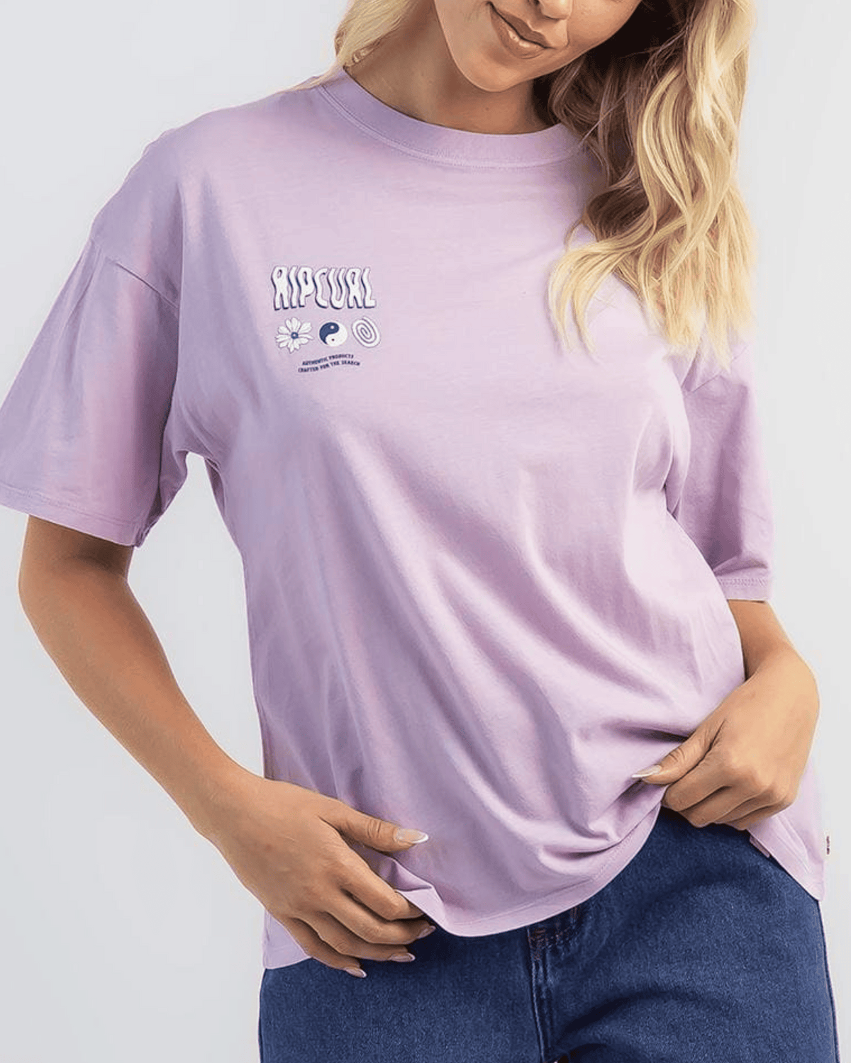A woman with wavy blonde hair exudes vintage charm in the Rip Curl Cosmic Dreams Heritage Tee, light purple with small chest designs and text, paired with blue jeans as she holds the hem of her shirt.