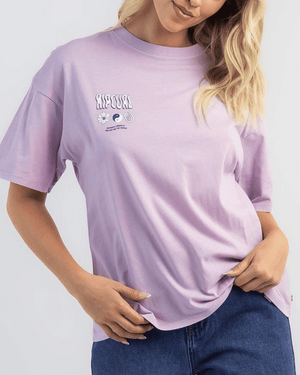 A woman with wavy blonde hair exudes vintage charm in the Rip Curl Cosmic Dreams Heritage Tee, light purple with small chest designs and text, paired with blue jeans as she holds the hem of her shirt.