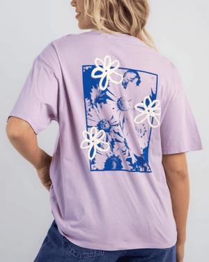 A woman stands with one hand on her hip, facing away, wearing a light purple Rip Curl Cosmic Dreams Heritage Tee featuring a blue and white floral graphic with three raised white flowers on the back, radiating vintage charm.