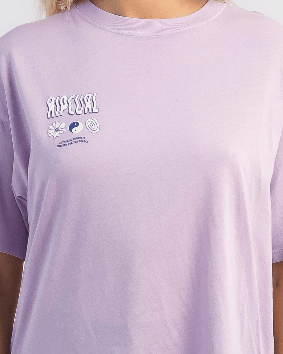 A person in a light purple Rip Curl Cosmic Dreams Heritage Tee with “RIP CURL” and small symbols—a flower, yin-yang, and spiral—on the upper left chest. The vintage-inspired heritage fit adds charm. The person's head is not visible.