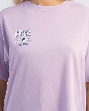A person in a light purple Rip Curl Cosmic Dreams Heritage Tee with “RIP CURL” and small symbols—a flower, yin-yang, and spiral—on the upper left chest. The vintage-inspired heritage fit adds charm. The person's head is not visible.
