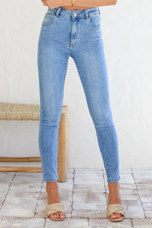 A woman stands on a tiled floor, wearing the Country Denim Ankle Length Stretchy Jean in light blue with woven beige sandals; a woven bench and light wall are seen in the background. Her upper body is mostly out of frame.