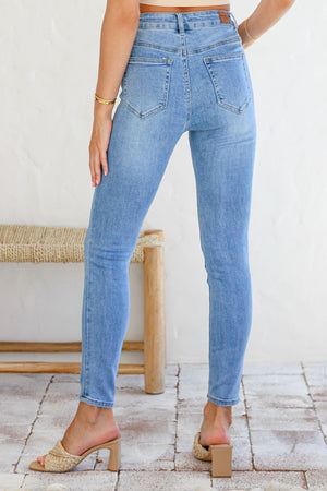 A person stands indoors facing away, wearing the light blue Country Denim Ankle Length Stretchy Jean and beige heeled sandals, with one hand on their hip. A woven bench and neutral-toned tiled floor complete the scene.