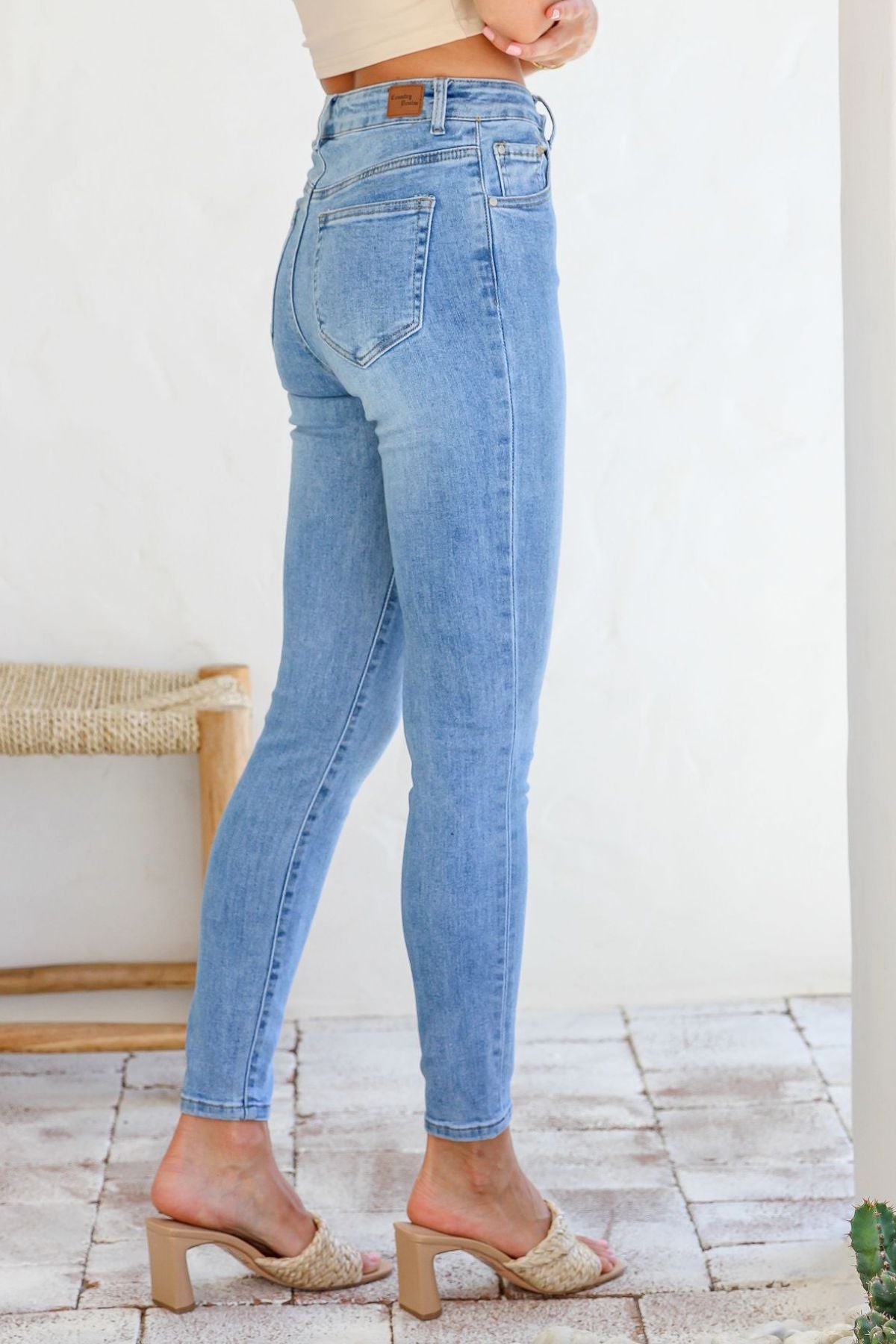A person models the Country Denim Ankle Length Stretchy Jean in light blue, standing sideways on a tiled floor with a woven bench and white wall behind them.