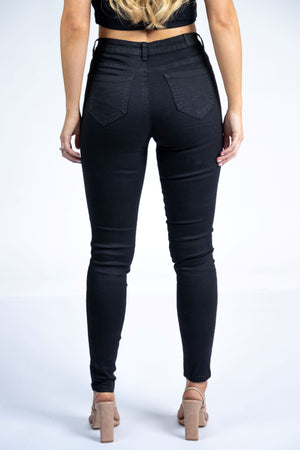 A woman faces away from the camera, wearing Country Denim Skinny Jeans, a black crop top, and beige high heels against a plain white background.