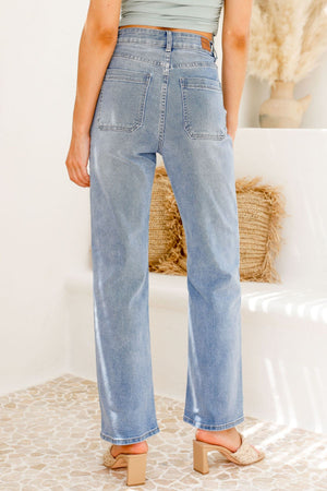 A person wearing Country Denim Straight Leg Stretchy Jean in light blue and block-heeled sandals stands indoors on a tiled floor with their back to the camera; a woven cushion and vase appear in the background.